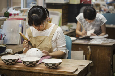 Woman working in a Japanese porcelain workshop, painting geometric pattern onto white bowls with paintbrush. - MINF07713