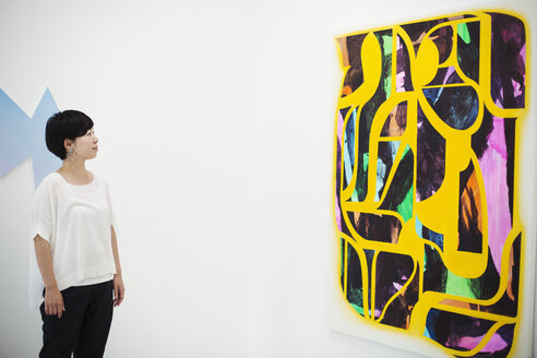 Frau mit kurzen schwarzen Haaren und weißem Hemd steht in einer Kunstgalerie und betrachtet ein modernes Gemälde. - MINF07715