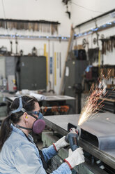 Frau mit Schutzbrille und Staubmaske steht in einer Metallwerkstatt und benutzt eine Schleifmaschine, wobei Funken fliegen. - MINF07761