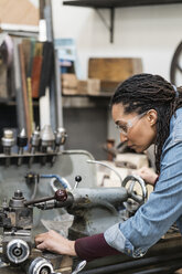 Frau mit Schutzbrille steht in einer Metallwerkstatt und arbeitet an einer Maschine. - MINF07766