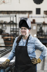 Porträt einer Frau mit Schürze und Schweißmaske in einer Metallwerkstatt, die in die Kamera lächelt. - MINF07768