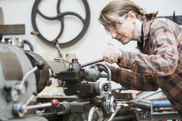 Eine Frau mit Schutzbrille steht in einer Metallwerkstatt und arbeitet an einer Drehbank. - MINF07772