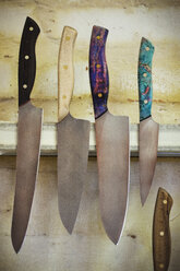Eine Auswahl von Messern mit geformten und bemalten Holzgriffen auf einem magnetischen Messerhalter an der Wand einer Werkstatt. - MINF07791