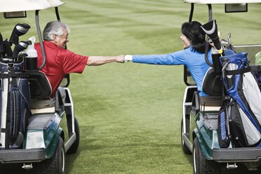 A senior couple doing a fist bump between golf carts on the fairway of a golf course. - MINF07837