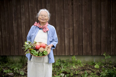 Elderly woman with grey hair standing in a garden, holding a basket with fresh vegetables, smiling at camera. - MINF07869