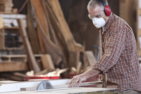 Ein weißer Fabrikarbeiter trägt einen Gehörschutz und eine Staubmaske für die Nase, während er mit einer Tischsäge Holz schneidet. - MINF07943