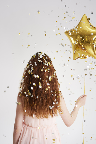 Rückenansicht einer rothaarigen jungen Frau mit einem sternförmigen goldenen Luftballon unter einem Konfettiregen, lizenzfreies Stockfoto