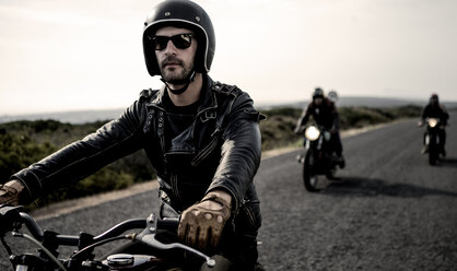 Ein Mann mit offenem Sturzhelm und Sonnenbrille fährt mit einem Cafe Racer Motorrad eine Landstraße entlang. - MINF07956
