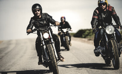 Drei Männer mit offenen Sturzhelmen und Sonnenbrillen fahren mit Cafe-Racer-Motorrädern eine Landstraße entlang. - MINF07957