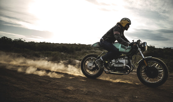Seitenansicht eines Mannes mit Sturzhelm auf einem Cafe-Racer-Motorrad auf einem staubigen Feldweg. - MINF07968
