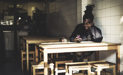 Mann mit Dreadlocks sitzt drinnen an einem Tisch, schaut auf sein Smartphone, Bierflasche auf dem Tisch. - MINF07972