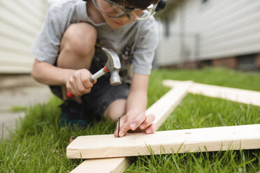 Young boy with brown hair kneeling in a garden, holding hammer and nail, hammering two wooden planks together. - MINF07982