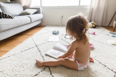 Hochformatige Seitenansicht eines jungen Mädchens in Unterhosen, das auf einem Teppich sitzt und ein Buch betrachtet. - MINF07993