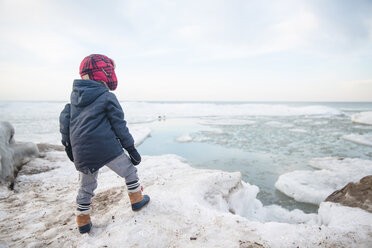 Rear view of boy wearing warm clothes standing on an icy ledge of a partially frozen lake. - MINF07996