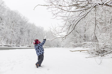 Junge mit Pelzmütze steht mit erhobenem Arm an einem von Bäumen gesäumten Fluss im Schnee. - MINF08004