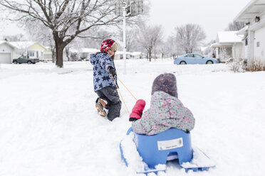 Boy wearing furry hat pulling young girl on a sledge cross snow-covered front lawn. - MINF08007