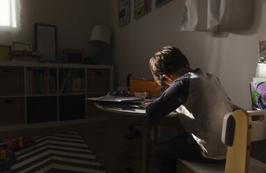 Rückansicht eines Jungen mit braunem Haar, der in einem Haus auf einem Stuhl an einem Tisch sitzt und schreibt. - MINF08011