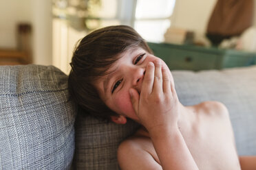 Junge mit braunen Haaren sitzt drinnen auf einem Sofa, hält sich den Mund mit der Hand zu und schaut in die Kamera. - MINF08019