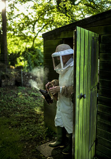 A beekeeper walking out of a shed wearing a veil and holding a smoker to calm honeybees. - MINF08064