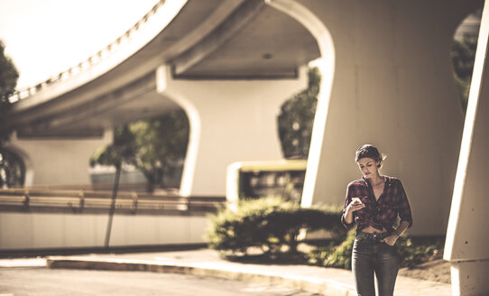 A woman walking under a road bridge looking at a mobile phone. - MINF08096