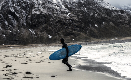 Ein Surfer im Neoprenanzug und mit einem Surfbrett in der Hand steigt aus dem Ozean, die Berge im Rücken. - MINF08109