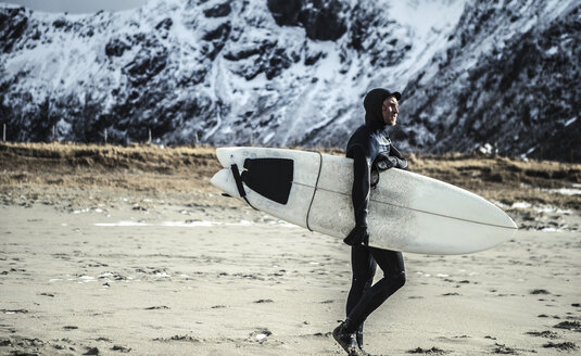 Ein Surfer in einem Neoprenanzug und mit einem Surfbrett auf dem Rücken läuft über einen Strand. - MINF08110