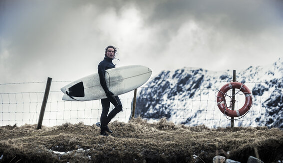 Ein Surfer in einem Neoprenanzug, der ein Surfbrett trägt und an einem Rettungsring vorbeigeht. - MINF08111