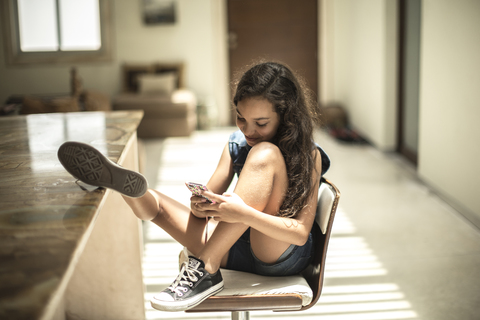 A girl sitting looking at a mobile phone screen with feet up on a kitchen counter. stock photo
