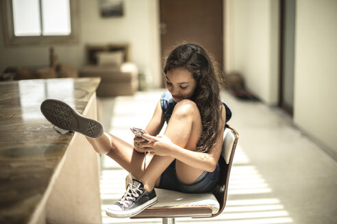 A girl sitting looking at a mobile phone screen with feet up on a kitchen counter. - MINF08120