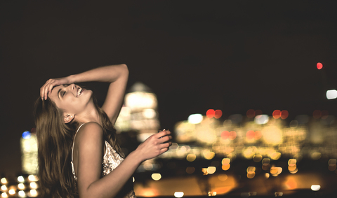 A young woman in a sequined party dress sitting on a rooftop at night with her head back and a drink in her hand. stock photo