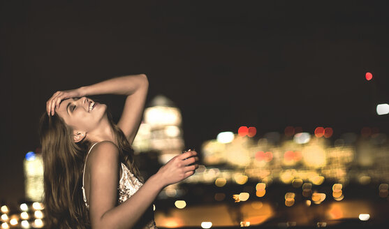 Eine junge Frau in einem paillettenbesetzten Partykleid sitzt nachts auf einem Dach, den Kopf in den Nacken gelegt und einen Drink in der Hand. - MINF08178