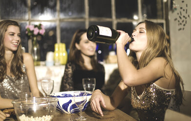 Eine Gruppe von Freunden sitzt auf einer Party um einen Tisch herum, ein Mädchen trinkt aus einer Sektflasche. - MINF08186