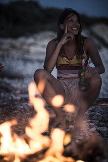 Eine junge Frau sitzt an einem Strand mit einem Lagerfeuer im Vordergrund. - MINF08191