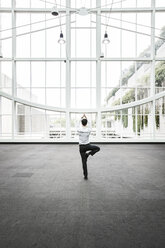 Ein Geschäftsmann entspannt sich bei einer Yogastellung in einem großen, offenen, glasüberdachten Gang. - MINF08205
