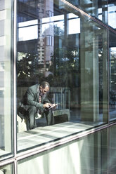 Black business man texting while sitting in front of a large widow in a lobby waiting area. - MINF08278