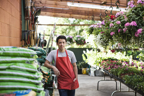 Junger Angestellter in roter Schürze, der sich in einer Gärtnerei an Säcke mit Kompost lehnt. - MINF08316