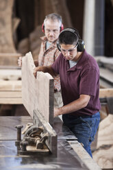 Team of two Caucasian men cutting a large wooden slab on a table saw in a woodworking factory. - MINF08370