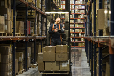 Warehouse worker wearing a safety harness while operating a motorized stock picker in an aisle between large racks of cardboard boxes holding product on pallets in a large distribution warehouse. - MINF08392