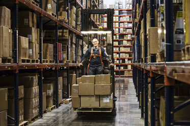 Warehouse worker wearing a safety harness while operating a motorized stock picker in an aisle between large racks of cardboard boxes holding product on pallets in a large distribution warehouse. - MINF08393