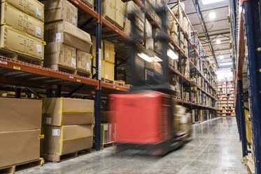 Blur of a motorized stock picker between aisles of cardboard boxes on pallets stacked on large racks in a large distribution warehouse. - MINF08395