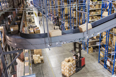 Overhead view looking down an aisle of large racks, conveyor belts and fork lifts, in a distribution warehouse of cardboard boxes holding products. - MINF08396