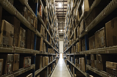 View down an aisle of racks holding cardboard boxes of product on pallets in a large distribution warehouse - MINF08398