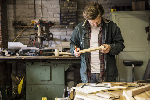 Mann arbeitet stehend an einer Werkbank in einer Holzwerkstatt und hält ein Stück Holz. - MINF08402