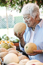 Eine ältere Frau steht an einem Bauernhofstand und hält sich eine frische Melone an die Nase, um daran zu riechen. - MINF08426