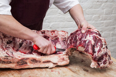 Close up of butcher wearing apron standing at a wooden butcher's block, butchering beef forerib. - MINF08429