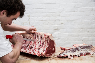 Close up of man wearing apron standing at a wooden butcher's block, butchering beef forerib. - MINF08430