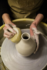 Nahaufnahme eines Töpfers mit Schürze bei der Arbeit an der Töpferscheibe, der eine Vase aus Ton formt. - MINF08435