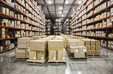 View down aisles of racks holding cardboard boxes of product on pallets in a large distribution warehouse - MINF08443