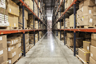 View down an aisle of racks holding cardboard boxes of product on pallets in a large distribution warehouse - MINF08444