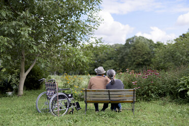 Ehemann und Ehefrau, Rückansicht eines älteren Mannes mit Hut und einer Frau, die nebeneinander auf einer Bank in einem Garten sitzen, neben der ein Rollstuhl geparkt ist. - MINF08449